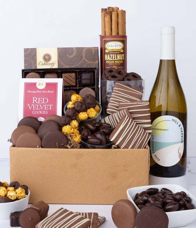 A gift box overflowing with treats including an assortment of chocolate-covered items like oreoes, almonds graham crackers and pretzels along with red velvet cookies, hazelnut wafers, an 8 piece box of chocolates and a bottle of white wine.