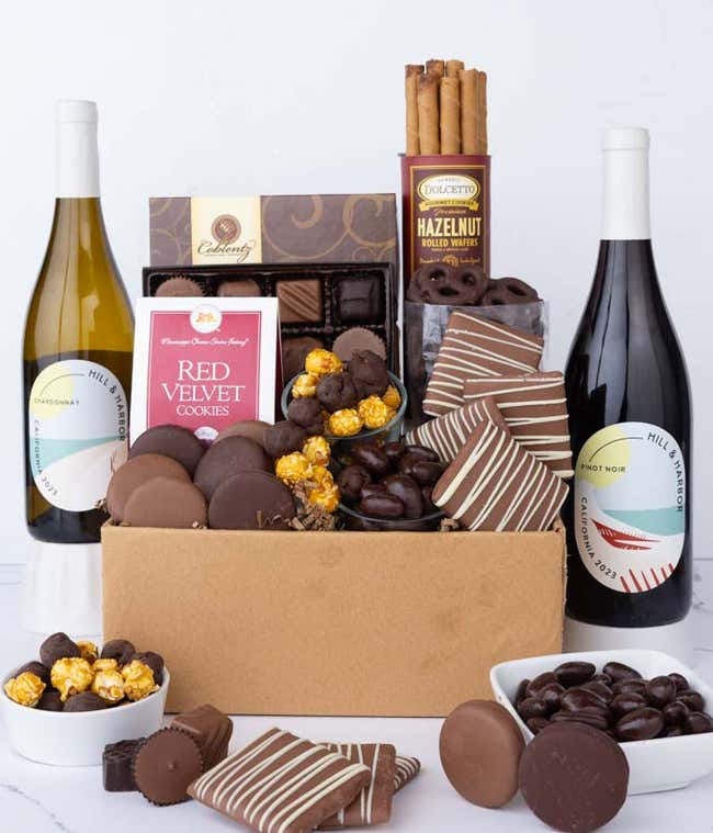 A gift box overflowing with treats including an assortment of chocolate-covered items like oreoes, almonds graham crackers and pretzels along with red velvet cookies, hazelnut wafers, an 8 piece box of chocolates and 2 bottles of wine.