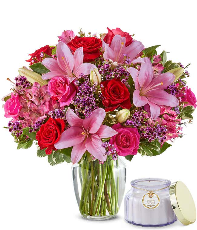 A bouquet in a glass vase of pink and red roses, pink lilies, pink and purple filler flowers sitting next to a small purple candle in glass jar. 