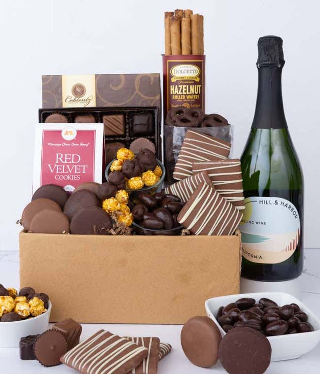 A gift box overflowing with treats including an assortment of chocolate-covered items like oreoes, almonds graham crackers and pretzels along with red velvet cookies, hazelnut wafers, an 8 piece box of chocolates and a bottle of sparkling wine.