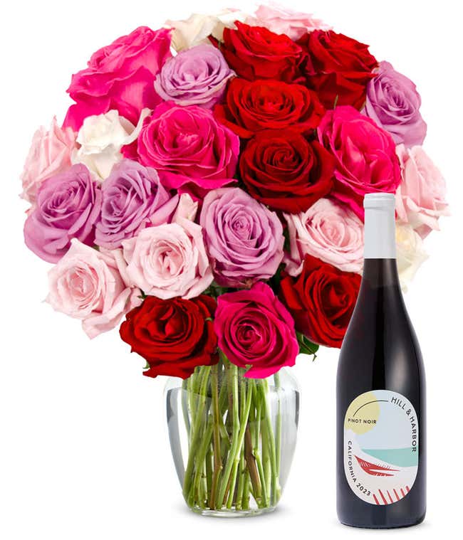 Two dozen pink, purple and red roses arranged in a clear glass vase sits beside a bottle of Hill &amp; Harbor California Pinot Noir.