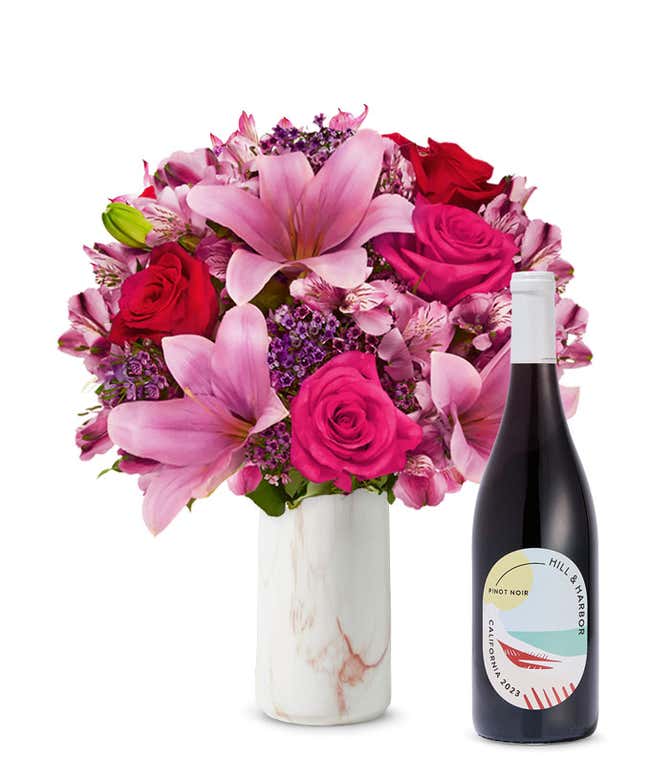 A bouquet of pink lilies, pink roses, and greenery arranged in a clear glass vase sitting next to a bottle of Hill &amp; Harbor California Pinot Noir.