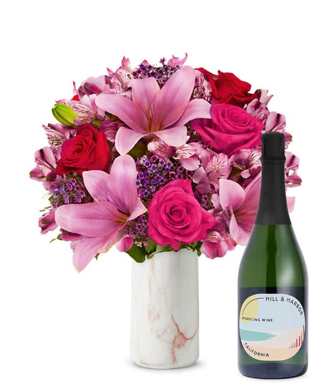 A bouquet of pink lilies, pink roses, and greenery arranged in a clear glass vase sitting next to a bottle of Hill &amp; Harbor California Sparkling Wine.