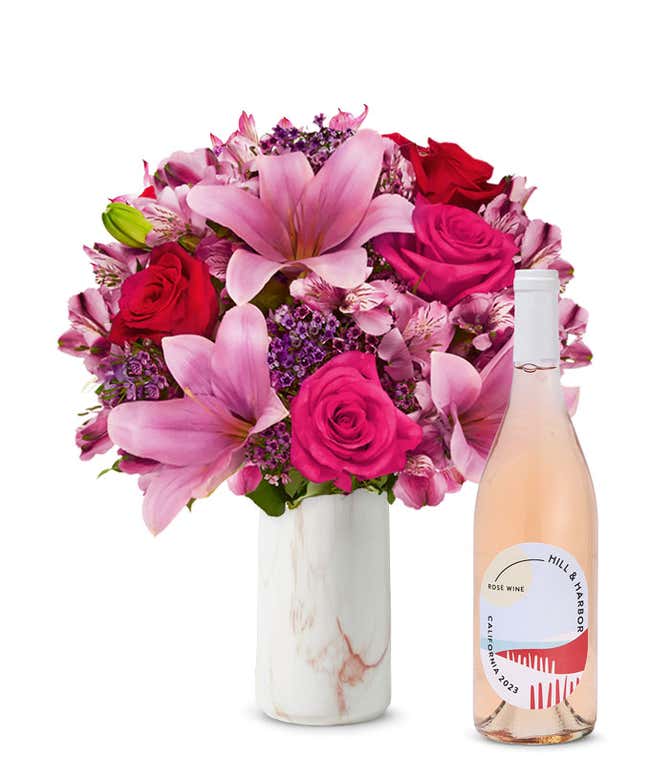 A bouquet of pink lilies, pink roses, and greenery arranged in a clear glass vase sitting next to a bottle of Hill &amp; Harbor California Ros&eacute;.