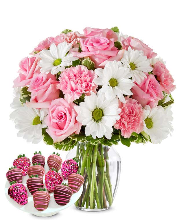 Deluxe bouquet of pink roses, pink carnations, and white daisy poms in a clear glass vase, paired with chocolate-covered strawberries decorated with pink drizzle and pink sprinkles.