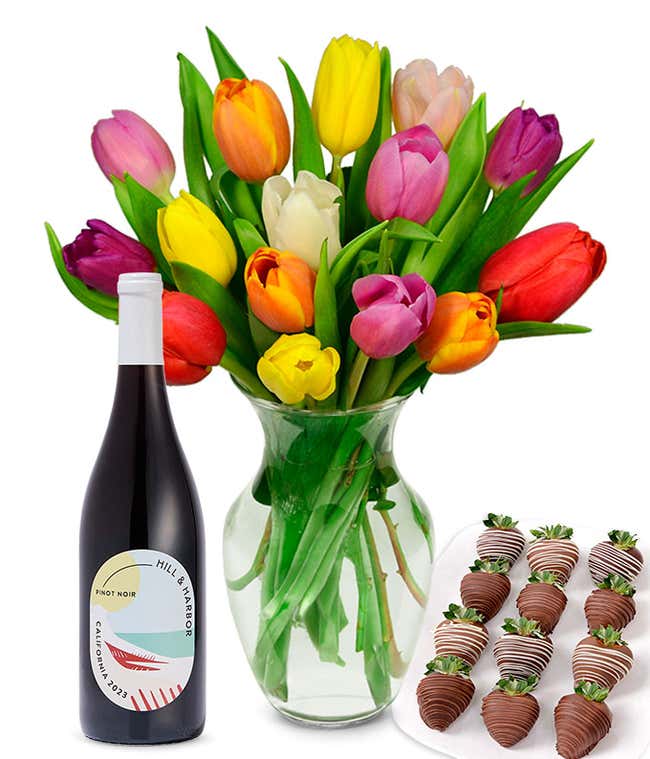 A clear glass vase filled with a 15-stem assortment of multi-colored tulips paired with a bottle of red wine and 12 chocolate-covered strawberries.