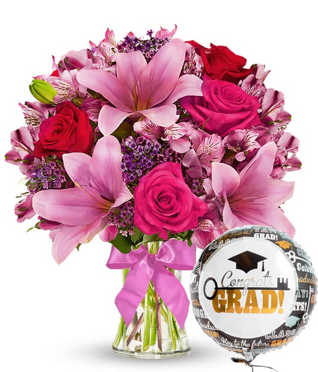A bursting full bouquet of red and pink flowers including roses, lilies, and more. Next to a congratulations mylar balloon. 