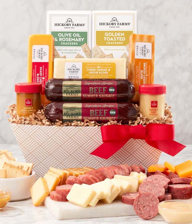 Gift basket with summer sausages, cheeses, crackers, and mustards in a white patterned container with a red bow. A serving board in front displays sliced sausage, cheese, and mustard for dipping.