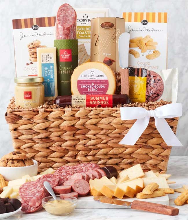 Woven gift basket with salami, cheese, mustard, crackers, cookies, and chocolates. In front, a board displays sliced meats, cheese cubes, and mustard for dipping, with cookies and chocolate truffles arranged alongside. Decorated with a white bow.