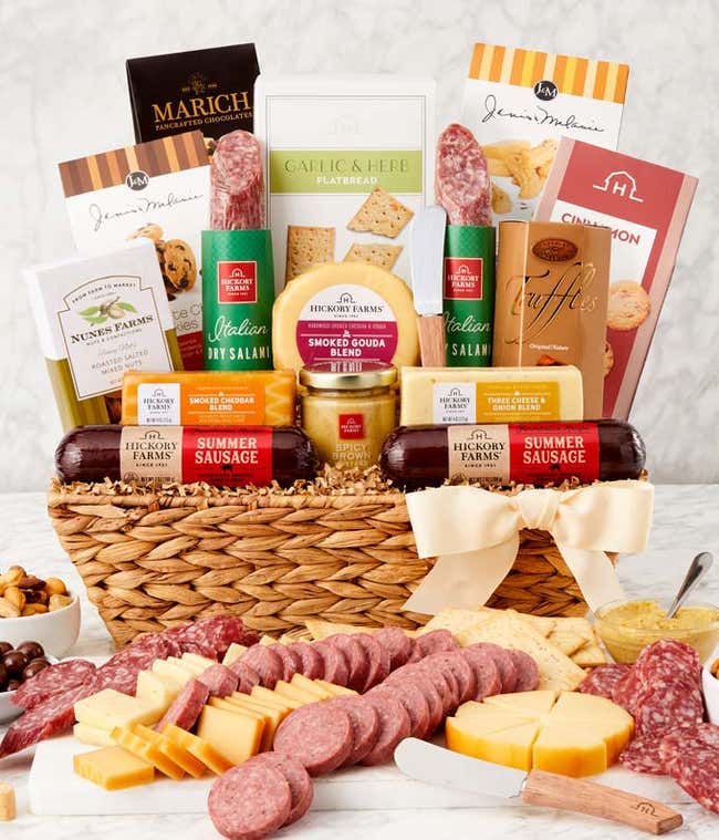 Woven gift basket filled with summer sausages, salami, assorted cheeses, crackers, mustards, cookies, chocolate truffles, and snacks. A serving board in front displays sliced sausage, cheese wedges, mustard, and pretzels, with a white bow on the basket.