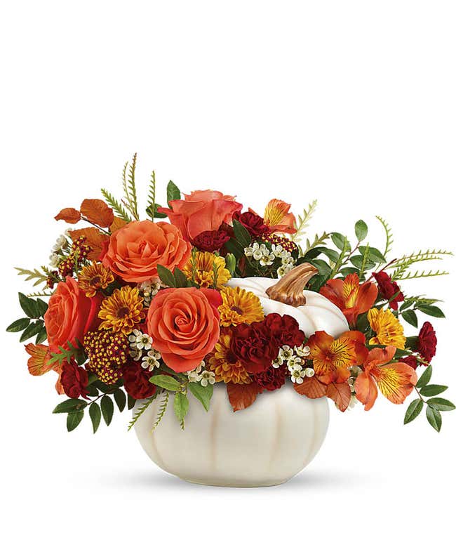 A charming floral arrangement featuring orange roses, orange alstroemeria, maroon mini carnations, yellow mums, and white waxflower, all beautifully arranged in a ceramic pumpkin.