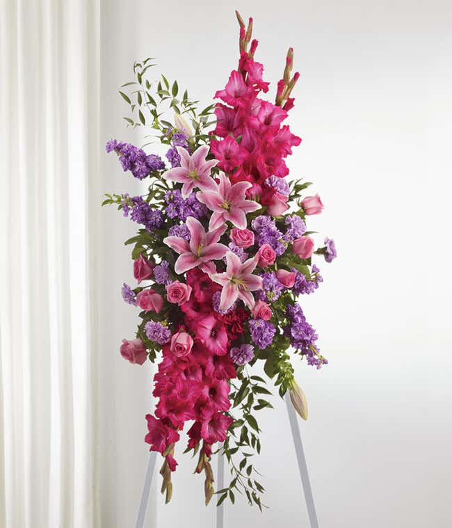Standing spray with pink gladiolus, lilies, roses, carnations, and green foliage on a white metal easel.