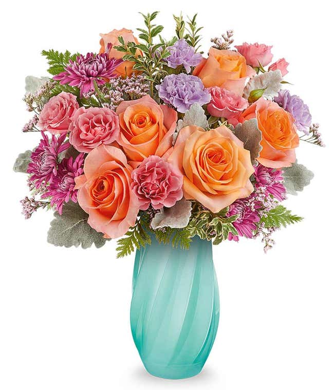  light orange roses, pink spray roses, lavender carnations, and purple poms in an enchanting aqua swirled vase with floral greenery.