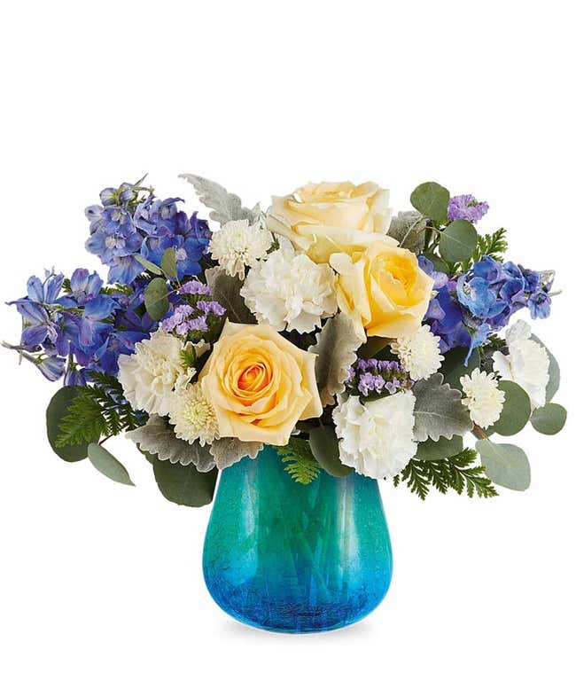 Floral arrangement with yellow roses, blue hydrangeas, white carnations, and greenery in a blue glass vase.