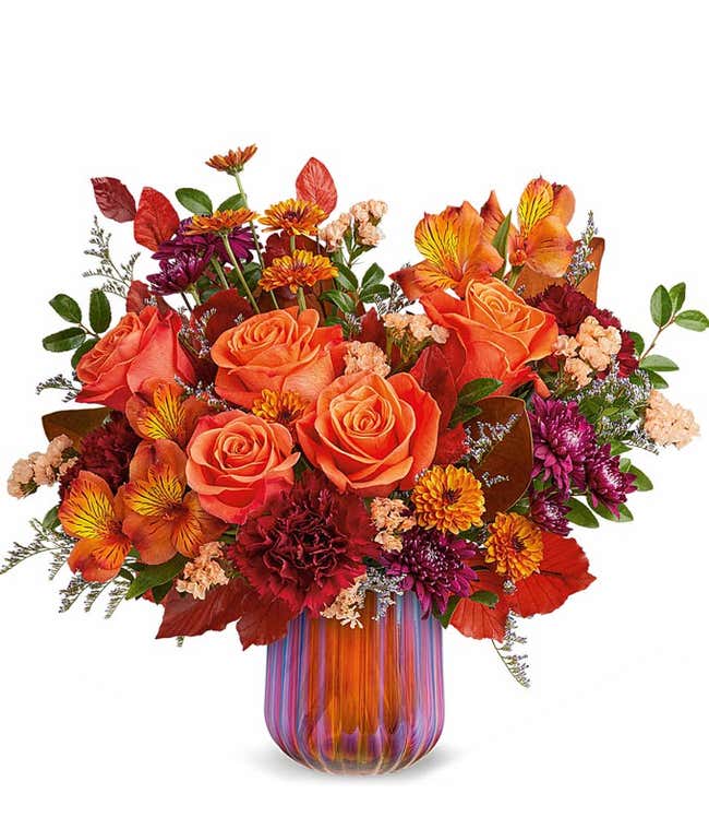 Orange Roses, Orange Alstroemeria, Burgundy Carnations, Bronze Poms, Purple Poms, and floral greenery in a sage green pitcher