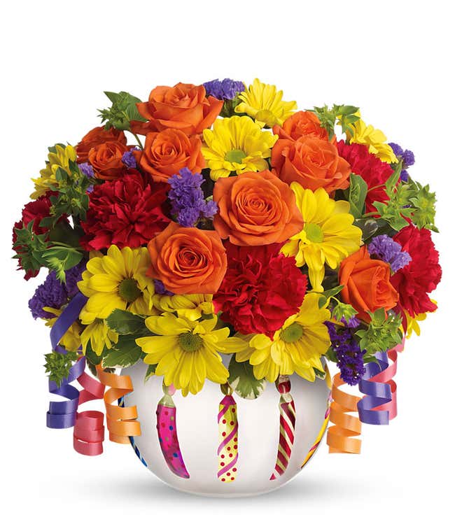 A festive arrangement featuring orange spray roses, red mini carnations, and yellow daisy spray mums, presented in a birthday cake inspired vase. This lively mix offers a fun and celebratory display.