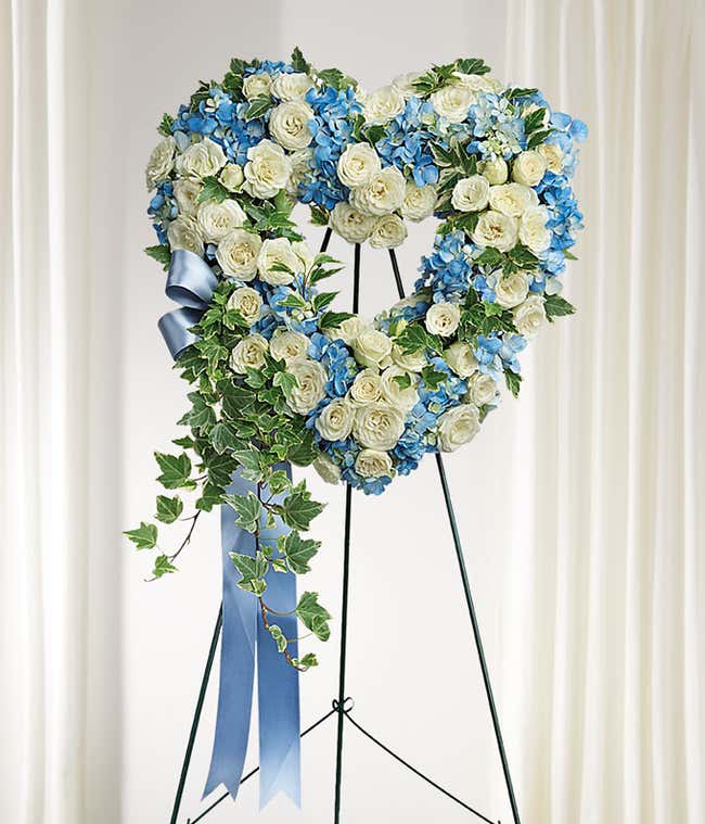 Heart-shaped funeral spray with white roses, blue hydrangeas, green ivy, and a blue ribbon on a metal easel.