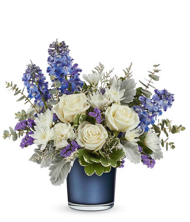 Elegant floral arrangement featuring white roses, white poms, deep blue delphinium, purple statice, dusty miller, silver dollar eucalyptus, and assorted greenery in a sleek navy blue glass vase.