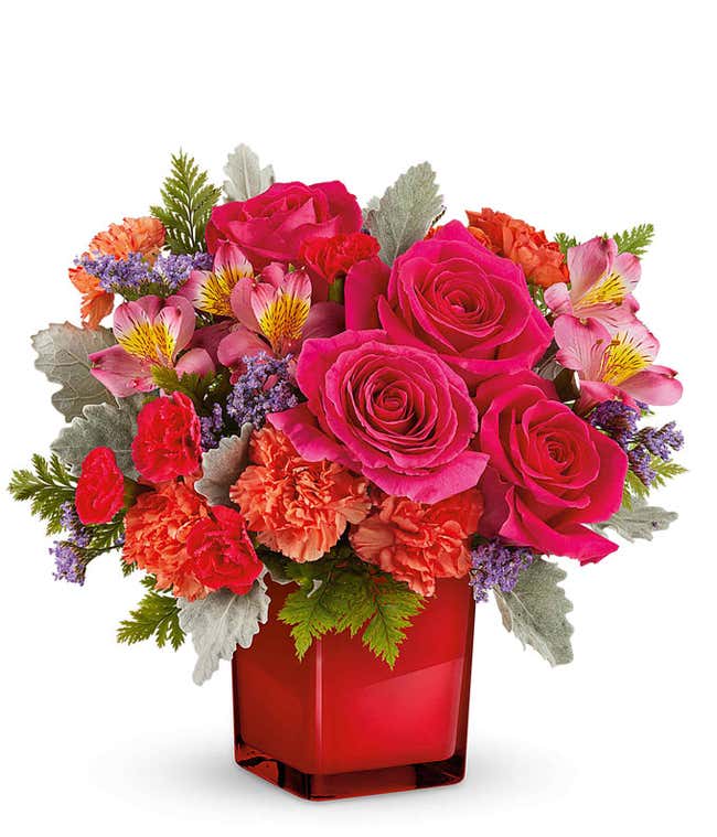 Floral arrangement with hot pink roses, pink alstroemeria, orange carnations, miniature red carnations, purple limonium, and assorted greenery in a red cube vase.