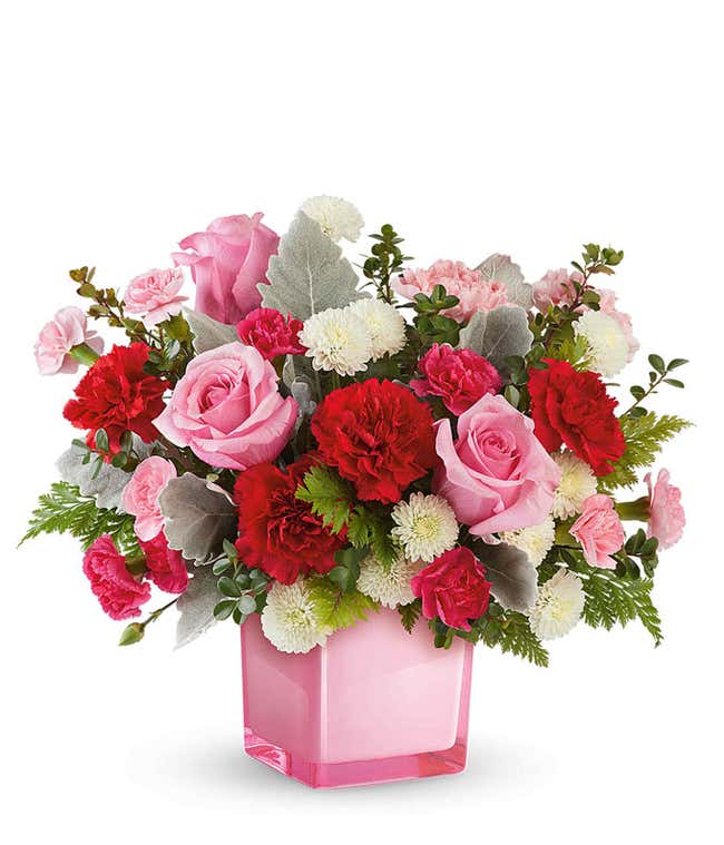 Pink and red roses with red and pink carnations, white mums, and greenery arranged in a soft pink glass cube vase