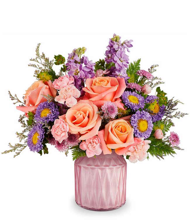 Peach roses, Mini pink carnations, lavender Matsumoto asters, lavender stock, and floral greenery in a pink glass vase with diamond texture