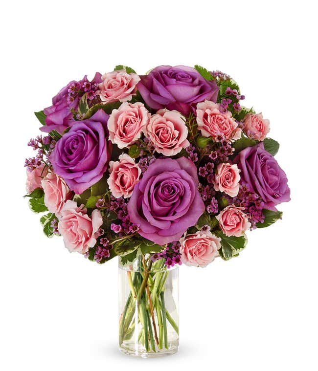 A vibrant bouquet of lavender roses, pink spray roses, and purple waxflower accented with lush greenery, arranged in a clear glass vase.