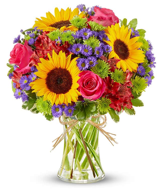 Bright mixed flower bouquet featuring yellow sunflowers, hot pink roses, red alstroemeria, green button poms, and purple asters in a clear glass vase tied with raffia ribbon.