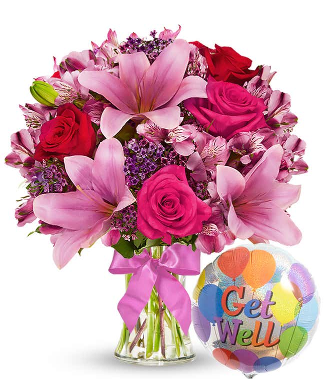 Clear glass vase filled with pink lilies, hot pink roses, red roses, pink alstroemeria, and purple filler flowers, tied with a pink bow and paired with a colorful 'Get Well' balloon. 