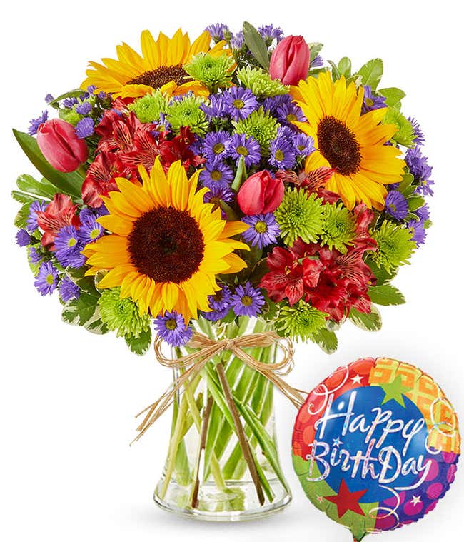 Clear glass vase filled with sunflowers, red alstroemeria, pink tulips, green button poms, and purple asters, tied with raffia and paired with a colorful Happy Birthday balloon.