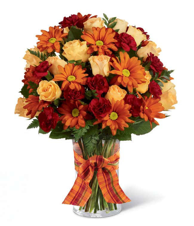 Clear glass vase filled with orange daisy poms, peach roses, and red mini carnations, accented with greenery and tied with an orange plaid ribbon.