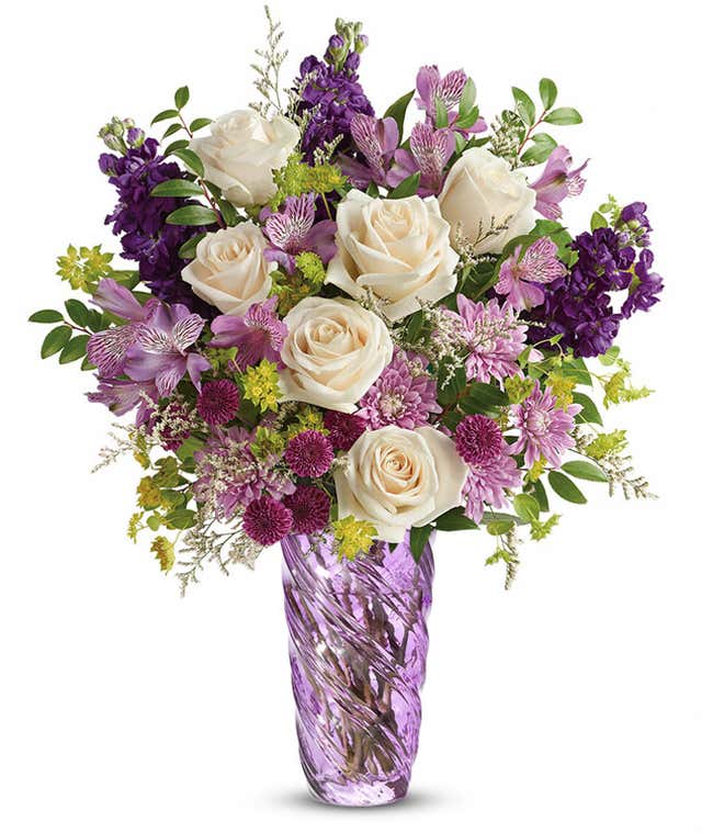 Floral arrangement featuring cr&egrave;me roses, lavender alstroemeria, purple stock, purple button chrysanthemums, lavender cushion spray chrysanthemums, white limonium, bupleurum, and huckleberry, all elegantly arranged in a lavender swirled vase.