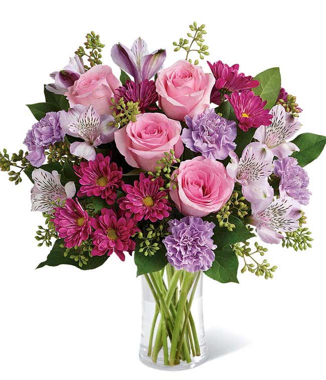 Pink roses, lavender carnations, magenta daisies, white alstroemeria, and lush greenery arranged in a clear glass vase, creating a soft and elegant bouquet in shades of pink and purple.