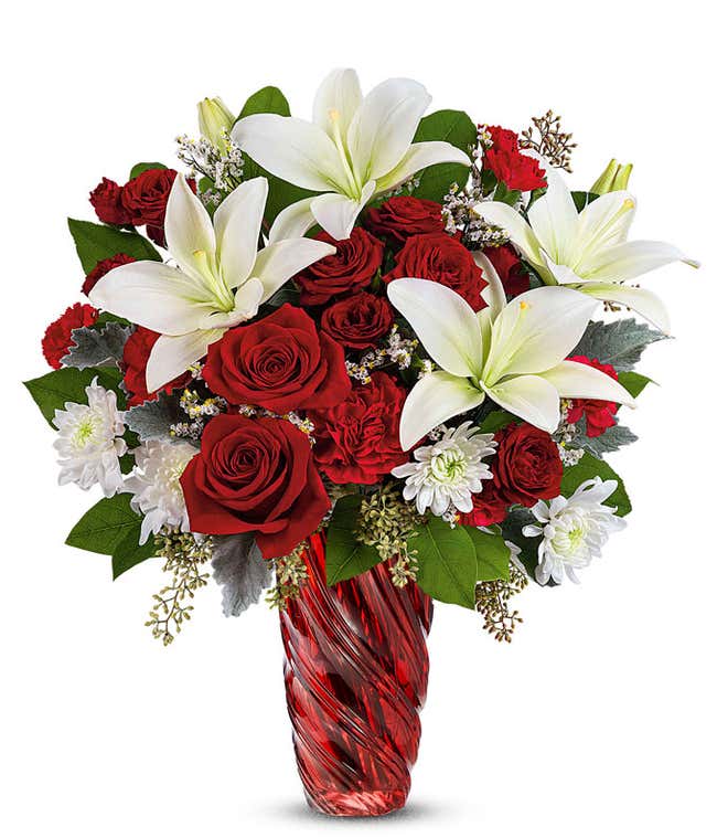 Red roses, white lilies, white poms, red carnations, and mixed greenery arranged in a red swirl glass vase.