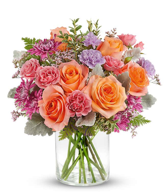 Vibrant floral arrangement featuring orange roses, pink miniature carnations, lavender carnations, pink cushion spray chrysanthemums, dusty miller, leatherleaf fern, and pink statice in a short glass cylinder