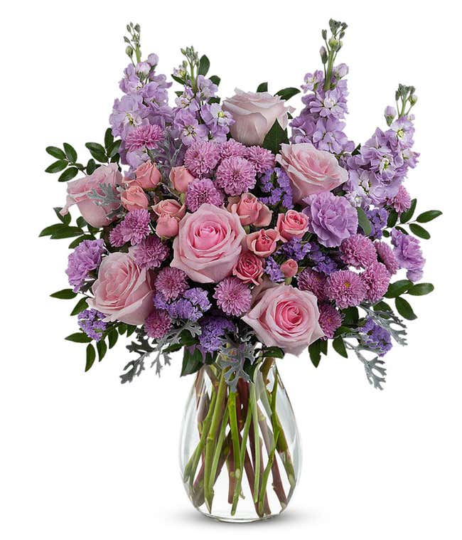 Pink roses, pink spray roses, lavender carnations, lavender stock, lavender button spray chrysanthemums, lavender sinuata statice, and floral greenery arranged in a clear glass jordan vase.