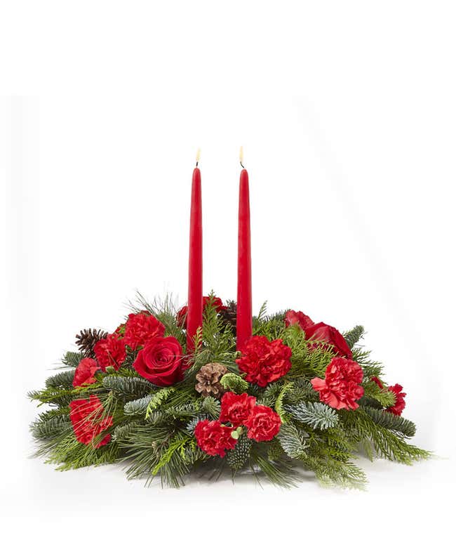 A festive arrangement with red roses, red carnations, red mini carnations, assorted seasonal greens, and red taper candles.