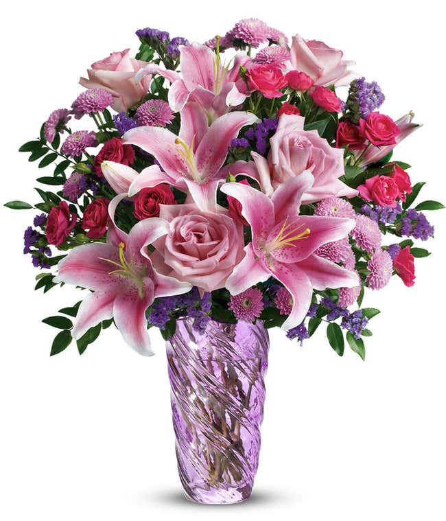 A bouquet of pink roses, stargazer lilies, hot pink spray roses, with purple and lavender filler flowers in a swirled lavender cylinder vase