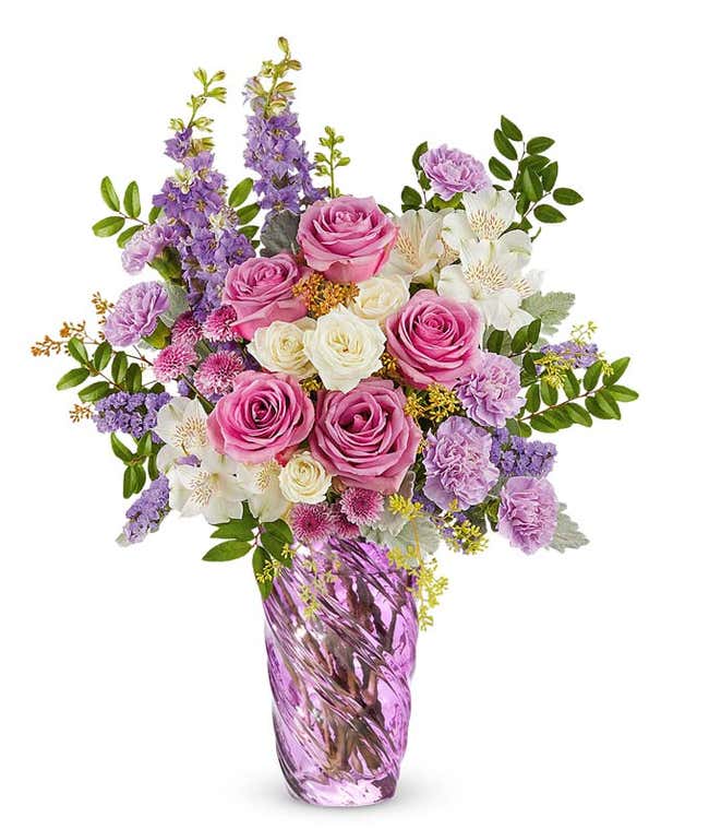 Lavender roses, white spray roses, white alstroemeria, lavender carnations, lavender larkspur, and lavender button poms with floral greenery in a swirled lavender vase
