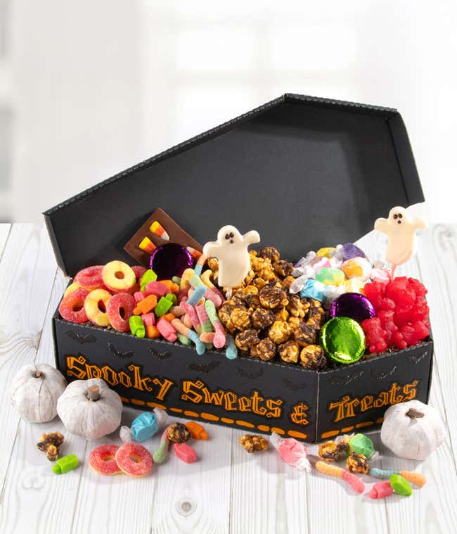 Halloween coffin-shaped gift box filled with chocolate-covered popcorn, ghost lollipops, gummy candies, foil-wrapped chocolates, salt water taffy, and assorted sweets, with decorative mini pumpkins and scattered candies around the box.