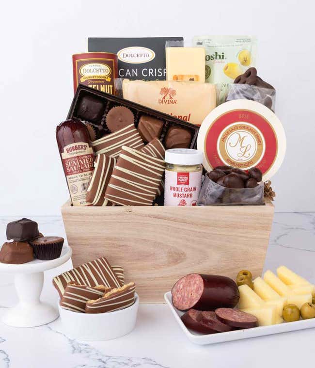 A wooden crate filled with gourmet snacks including sausage, assorted cheeses, crackers, olives, mustard, chocolate almonds, pretzels, graham crackers, assorted chocolates, and wafers, with plates of sausage, cheese, and chocolates in front.