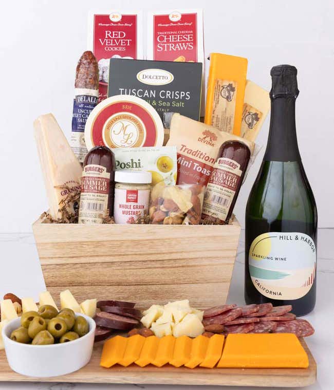 Wooden basket filled with 4 cheeses, 3 sausages, crackers, cookies, nuts, mustard, olives and crisps alongside a bottle of Hill &amp; Harbor Sparkling Wine.