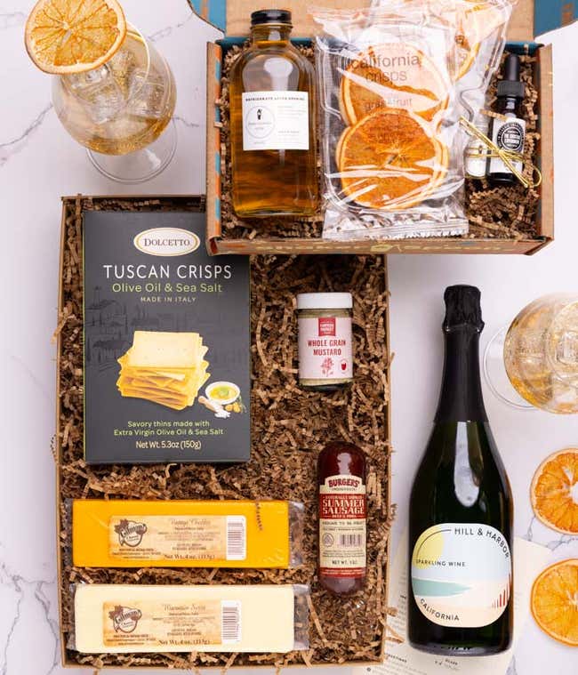 A bottle of Sparkling Wine next to a food box containing two blocks of cheese, summer sausage, crackers and mustard.  The cocktail box contains thyme-cucumber syrup, celery bitters, grapefruit oil mist, dehydrated grapefruit slices, and recipe card.