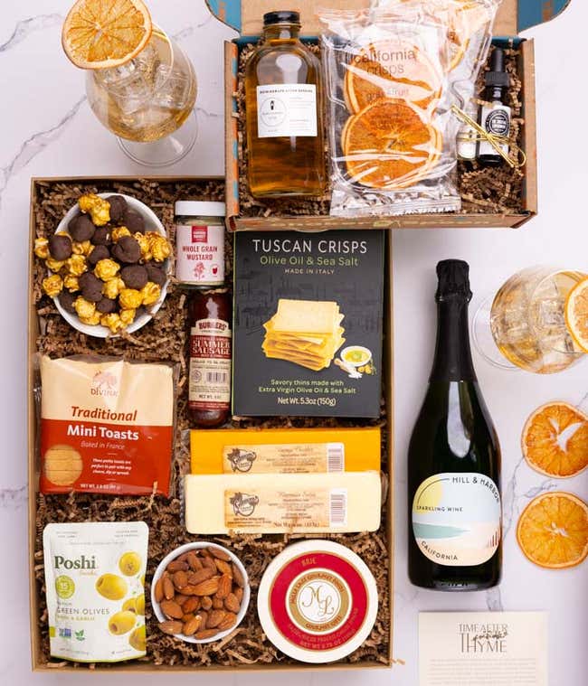A bottle of Sparkling Wine next to a box holding three cheeses, sausage, crackers, olives, almonds, popcorn &amp; mustard.  The cocktail box contains thyme-cucumber syrup, celery bitters, grapefruit oil mist, dehydrated grapefruit slices, &amp; recipe card.