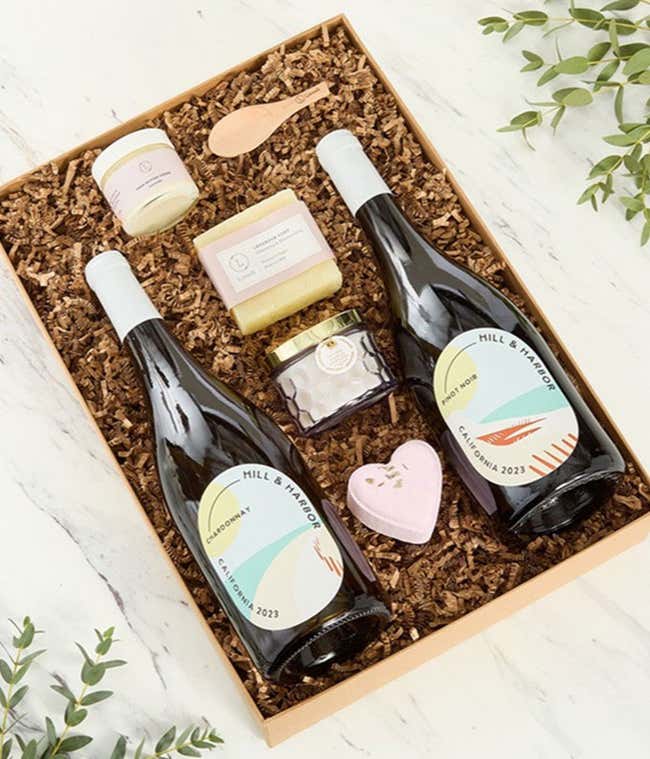 Two bottles of Hill &amp; Harbor wines displayed with spa essentials including artisan soap, bath salts with wooden scoop, and a scented candle arranged on a white surface, creating an elegant wine and self-care gift set.