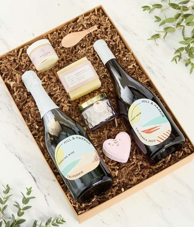 Two bottles of Hill &amp; Harbor wines displayed with spa essentials including artisan soap, bath salts with wooden scoop, and a scented candle arranged on a white surface, creating an elegant wine and self-care gift set.