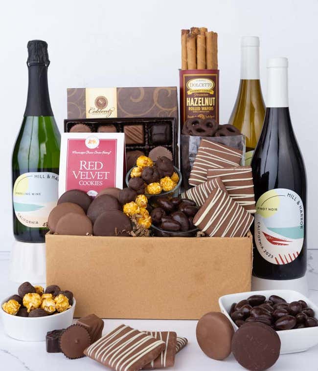 A gift box overflowing with treats including an assortment of chocolate-covered items like oreoes, almonds, graham crackers and pretzels along with red velvet cookies, hazelnut wafers, an 8 piece box of chocolates and 3 bottles of wine.