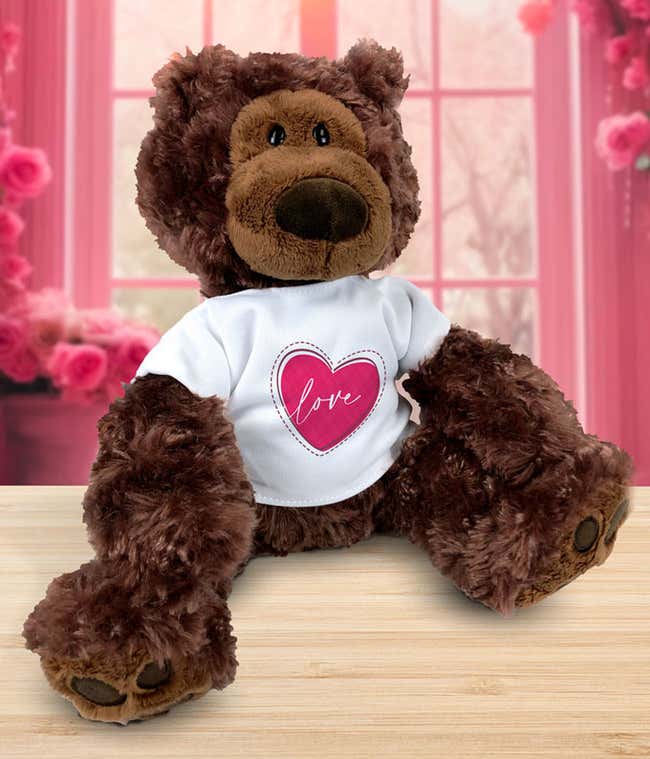 Plush brown teddy bear sitting on a wooden surface, wearing a white T-shirt with a pink heart design and the word love, set against a pink floral background.